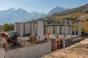 Construction workers build a concrete structure beside terraced hills, with snow-capped mountains in the background